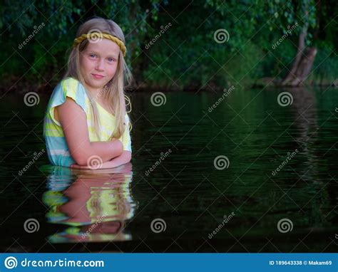Blonde Girls With Blue Eyes Bathes In Clothes In The Dark Water Of A Lake At Sunset Stock Photo