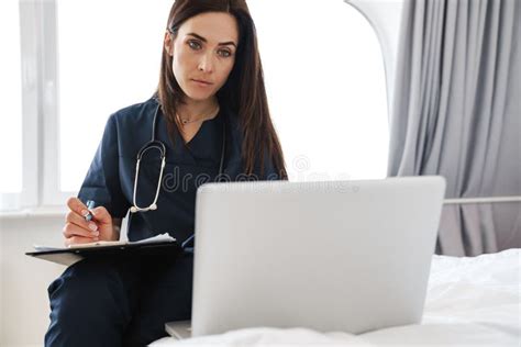 A Woman Doctor In A Medical Suit With A Stethoscope Sitting On The Bed