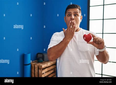 Hispanic young man holding red heart at gym covering mouth with hand ...