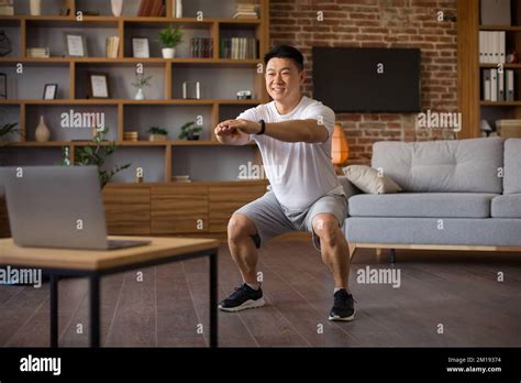 Athletic Korean Mature Man Doing Deep Squats During Online Workout At Home Looking At Laptop