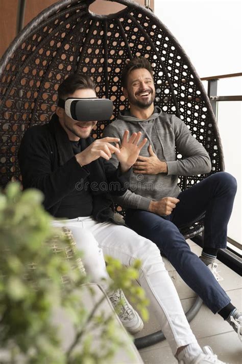 Amused Gay Couple Testing The Virtual Reality Glasses At Home Sitting In The Swing On The