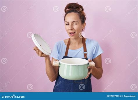 Brunette Woman Wearing Apron Holding Cooking Pot Winking Looking At The Camera With Expression