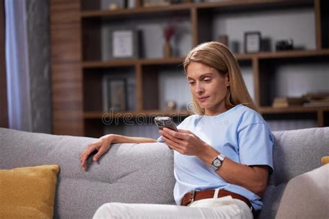 Blonde Girl Sitting On The Couch Looks Into The Smartphone Chatting Relaxing At Home Smiling