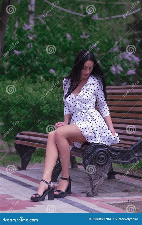 Beautiful Fashionable Brunette Woman Adult Sitting On A Bench The Park On The Plants Backgraund