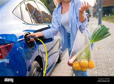 A Blonde Woman Plugs In Her Electric Vehicle As She Holds A Mesh Bag Filled With Fresh