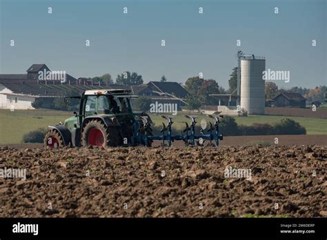 Tractor Ploughing A Field With Blurry Farm In The Background Stock
