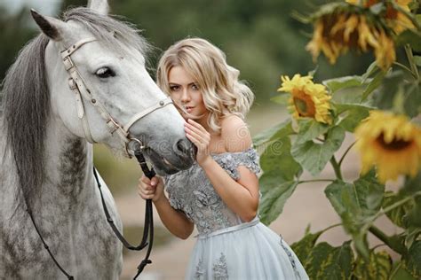 La Belle Fille Blonde Dans La Robe Frotte Un Cheval Gris Sur La Nature Dedans Photo Stock