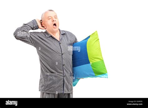 A Sleepy Mature Man In Pajamas Holding A Pillow And Yawning Isolated On