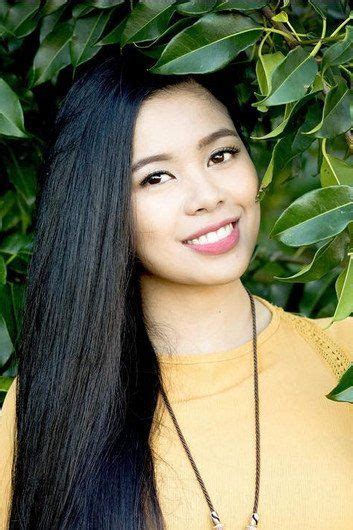 A Woman With Long Black Hair Standing In Front Of Green Leaves And Smiling At The Camera