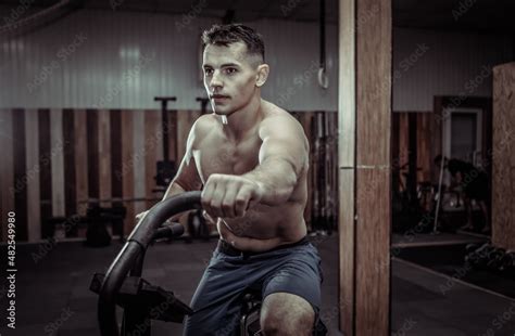 Powerful Muscular Male Athlete With A Naked Torso Trains With An Air Bike In A Modern Gym