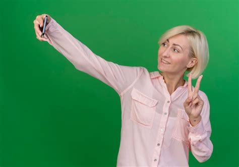 Lächelnde junge blonde slawische Frau Friedenszeichen tut und selfie lokalisiert auf grünem