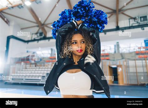 A Brunette Cheerleader With Her Arms Lifted High And Blue Pom Poms In Her Hands High Quality