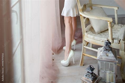 Slender Naked Female Legs In White Shoes A Woman In A Short Dress Stands Near The Window Stock