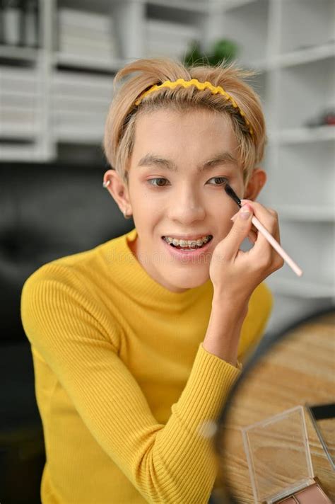 A Happy Asian Gay Man Is Looking At The Mirror And Applying Eyeshadow