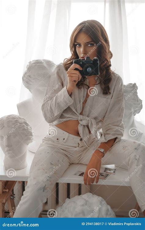 Elegant Brunette With Camera Seated Among Ancient Greek Busts On A White Table Stock Image