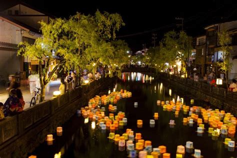 Many Candles Are Floating In The Water Near Buildings And People