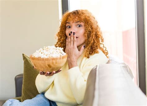 Mujer bonita latina de pelo rojo joven comiendo palomitas de maíz en un sofá y viendo una