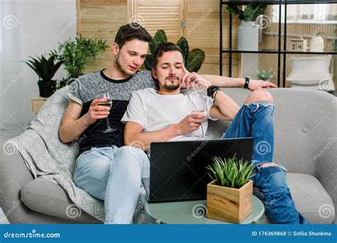 Happy Gay Couple Sitting On Sofa Watching Movie And Drinking Wine Gay Couple Homosexual