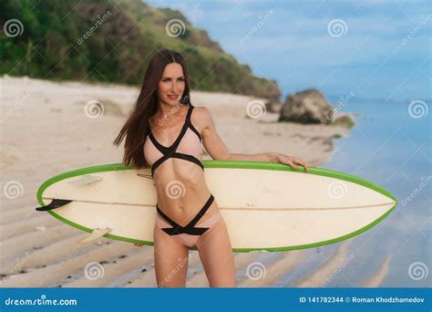 Beautiful Brunette Girl In Swimsuit Goes On Sandy Beach With Surfboard In Hand Stock Photo