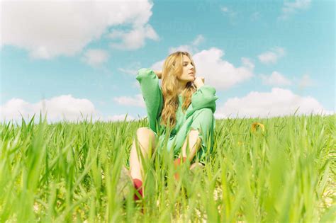 Blonde junge Frau in grünem Kleid sitzt auf einem Feld und schaut in Ferne lizenzfreies