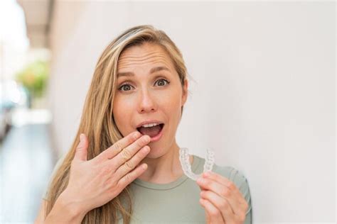 Junge blonde frau im freien hält unsichtbare zahnspangen mit überraschtem gesichtsausdruck