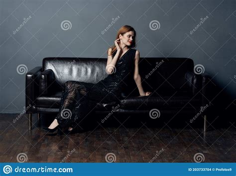 Woman In Black Dress Sits On Leather Sofa Indoors Evening Makeup Naked Shoulders Model Stock