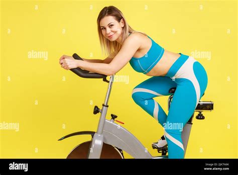 Side View Of Winsome Blonde Sporty Woman Riding Spinning Bike Looking At Camera With Positive