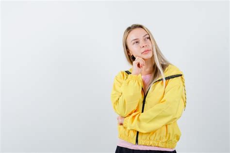 Fille blonde en veste jaune soutenant le menton sur place et à la pensive Photo Gratuite