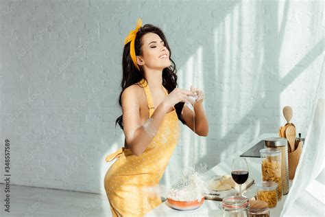 Smiling Nude Girl In Apron Adding Egg In Flour While Cooking On Kitchen Worktop Stock Photo