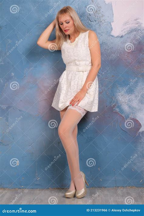 Blonde Legged Photomodel In White Minidress And White Stockings Posing In Studio Stock Photo