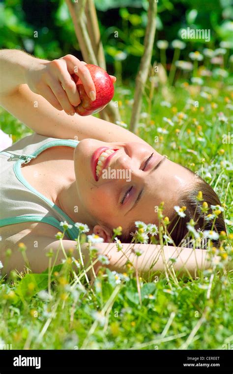 Brunette Female With Hair Slicked Back Lying In The Grass And Holding A Red Apple Above Head