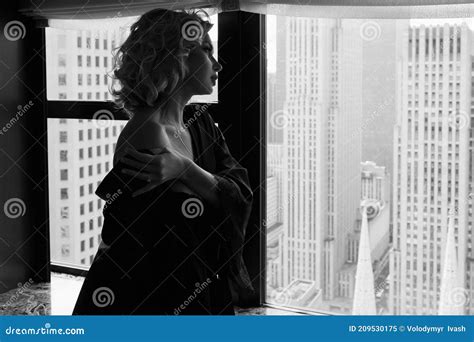 Woman In Black Silk Robe Stands With Naked Shoulders Before A Panoramic Window In New York Stock