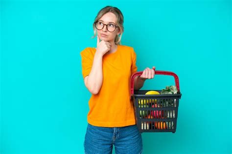 Blonde jeune fille anglaise tenant un panier plein de nourriture isolé sur fond bleu ayant des