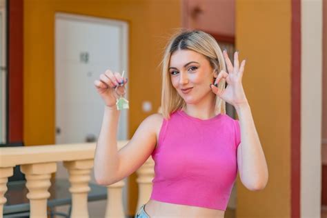 Junge hübsche blonde Frau mit Hausschlüsseln im Freien zeigt Ok Zeichen mit den Fingern