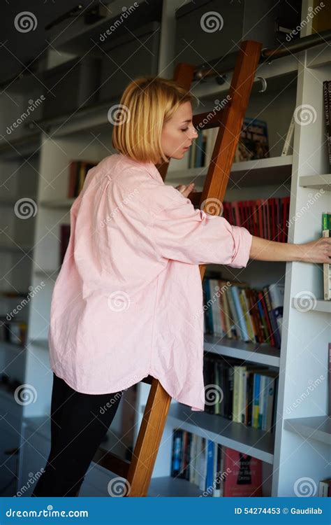 Charmante Blonde Haarvrouw Status Op Ladder Bij Boekenrek Terwijl Het Plukken Van Het Boek Stock