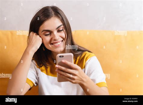 Cute Long Haired Brunette Girl Sitting On The Yellow Sofa In The Light Living Room And Browsing