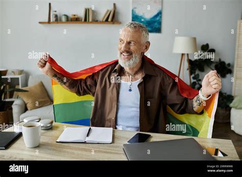 A Mature Gay Man With Tattoos And Grey Hair Proudly Holds A Rainbow Pride Flag In His Home Stock