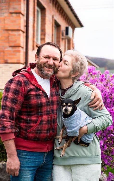 Mature Russian Married Couple Kissing And Hugging Near Blossom Bush Of Rhododendron Holding A