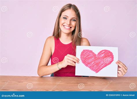 Jeune Fille Blonde Assise Sur La Table Tenant Le Coeur Dessiner Sourire Avec Un Sourire Joyeux