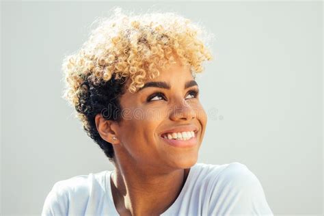 Hispanic Latin Woman With Afro Blonde Haircut Dressed In White Top Looking Up With Charming Shy