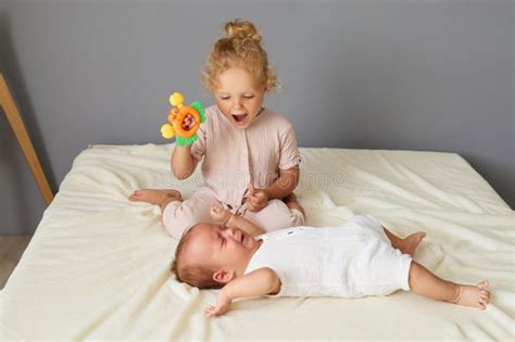 Older Sister With Blonde Hair Playing With Her Newborn Brother Showing Him Colorful Toy Indoors