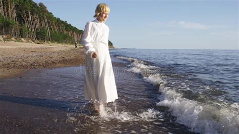 A Girl In A White Dress Walks Along The Seashore In The Rays Of The