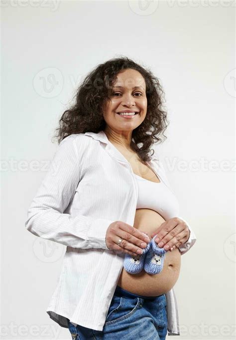 Beautiful Multi Ethnic Pregnant Woman Holding Blue Knitted Baby Booties On Her Naked Belly