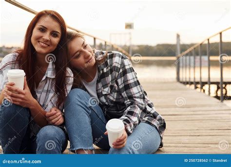 Cute Lesbian Couple Sits Together Near The Lake With Cups Of Drink In Hands And Embracing Each