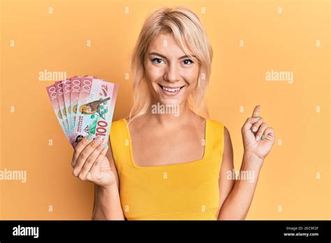 Jeune fille blonde tenant 100 dollars néo zélandais billet souriant pointez avec votre main et