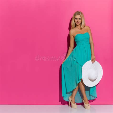 La Belle Femme Blonde Dans La Robe Et Des Talons Hauts De Turquoise Tient Le Chapeau Blanc De