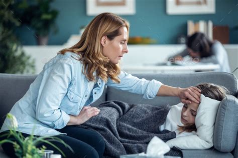 Mature Mother Caring For Her Sick Daughter While She Rests On The Sofa In Living Room At Home