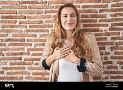 Beautiful Blonde Woman Standing Over Bricks Wall Smiling With Hands On Chest With Closed Eyes