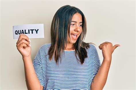 Jovem Latina Segurando A Palavra De Qualidade No Papel Apontando O Polegar Para O Lado Sorrindo