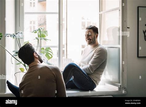 Happy Gay Man Talking To Boyfriend While Sitting On Windowsill At Home Stock Photo Alamy